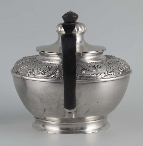 S/Mt/542/ML - Korpus w kształcie spłaszczonej kuli, na niskiej stopie, wylew w formie dzióbka, ucho drewniane malowane na czarno, osadzone w dwóch tulejkach, pokrywka na zawiasie, płaska, w części środkowej wypiętrzona i ozdobiona puklowaną rozetą.Górna część brzuśca ozdobiona sztancowaną taśmą z wzorem spiralnie zwinietej wici roślinnej z kwiatami, uchwyt przykrywki zakończony drewnianą rozetką.  
