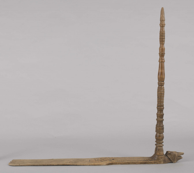 E/8019/ML - Przęślica krężołkowa z drewna brzozowego składa się z cienkiej deski – przysiadki, w której przy jednym końcu osadzona jest pionowa beleczka zw. piórem, z nasadzonym od góry ruchomym krężlem w kształcie wydłużonego stożka. Deseczka przysiadki o kształcie prostokątnym lekko zwężającym, od ok. połowy swej długości w kierunku pióra, i zakończonym głową zwierzęcą – psa. Pióro i krężel ozdobnie toczone. Krężel ma kształt stożka z rozszerzoną podstawą, w której znajduje się stożkowaty otwór, do osadzenia krężla na piórze. Dekoracja snycerska ryta znajduje się na zwężającej części przysiadki. Część zdobiona oddzielona małym stopniem. Grubość przysiadki w części zdobionej 2x większa od pozostałej części. Dekoracja snycerska tworzy ornament roślinny w postaci trzech ulistnionych drobno gałązek. Na zakończeniu ornamentu znajdują się ryte liście oraz inicjały „K.F.”. Głowa psa zdobiona drobnymi nacięcia. Zakończenie przysiadki wycięte faliście.