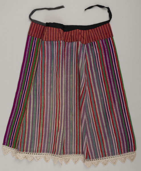 E/8132/ML - Fartuch wełniany szyty maszynowo. Wykonany z materiału samodziałowego tkanego w pionowe paski, w kolorach: czerwonym, białym, szafirowym, bordowym, zielonym. Uszyta z prostokątnego kawałka materiału, do którego doszyto z obu stron kliny. U góry fartuszek przedłużony przez wszycie kawałka materiału szer. 7 cm, w pasy czerwono-granatowe. W talii wszyty w pasek z czarnego materiału, szer. 2 cm, zakończony troczkami. Dół fartuszka zdobi biała koronka szer. 5 cm.