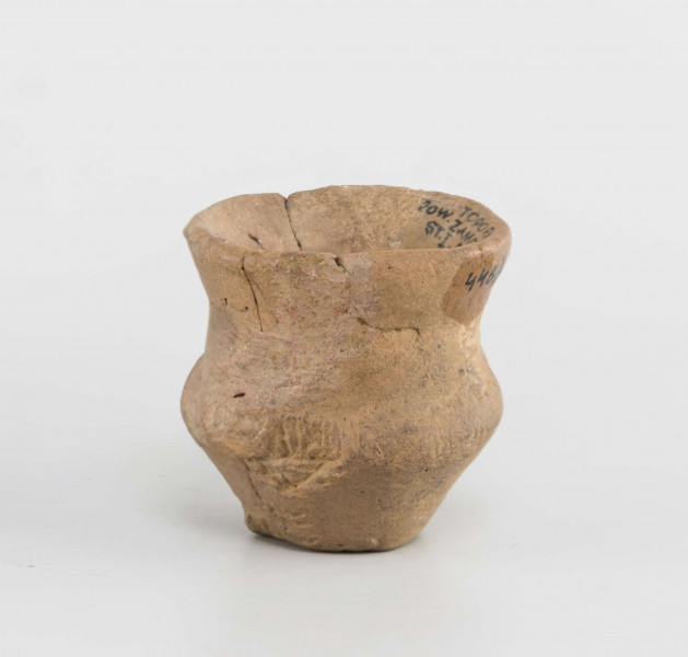 Naczynko miniaturowe. Dno małe, płaskie, wyodrębnione. Brzusiec silnie załamany. Szyjka wyodrębniona, wysoka. Krawędź zaokraglona, wychylona. Powierzchnia gładzona, na zewnętrznej odciski roślinne. Surowiec - glina z domieszką piasku. Barwa całości brunatno-pomarańczowa. Zabytek sklejany, drobne ubytki uzupełnione gipsem.