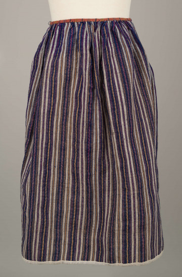 E/5997/ML - Fartuszek uszyty z jednego kawałka materiału - tkaniny samodziałowej bawełniano-wełnianej, tkanej splotem rypsowym w drobne paseczki w kolorze: białym, czerwonym, granatowym i bordo. Suto marszczony w oszewkę przechodzącą w troczki. Oszewka z materiału lnianego w kratkę czerwono-zielono-granatową. Dół obszyty tasiemką z białego bawełnianego płótna.