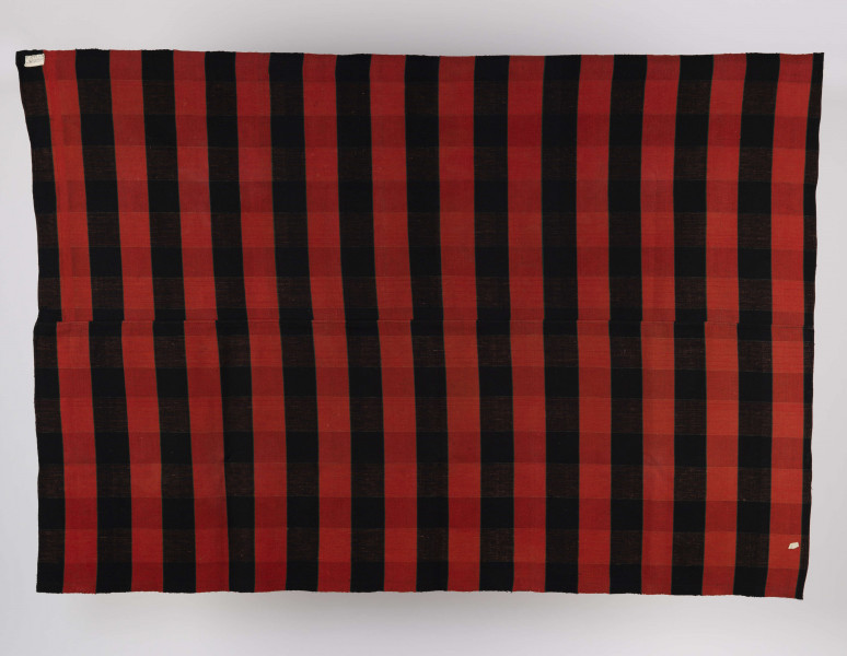 E/6550/ML - Tkanina kraciak tkana, zszyta z 2 części pośrodku. Splot rządkowym. Osnowa lniana czarna, wątek wełniany czerwony. Raport tworzą pasy o szer. 7,5 cm. Krzyżujące się nici zestawu osnowy i wątku dają lekko zaznaczoną kratę.