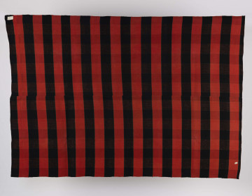 E/6550/ML - Tkanina kraciak tkana, zszyta z 2 części pośrodku. Splot rządkowym. Osnowa lniana czarna, wątek wełniany czerwony. Raport tworzą pasy o szer. 7,5 cm. Krzyżujące się nici zestawu osnowy i wątku dają lekko zaznaczoną kratę.