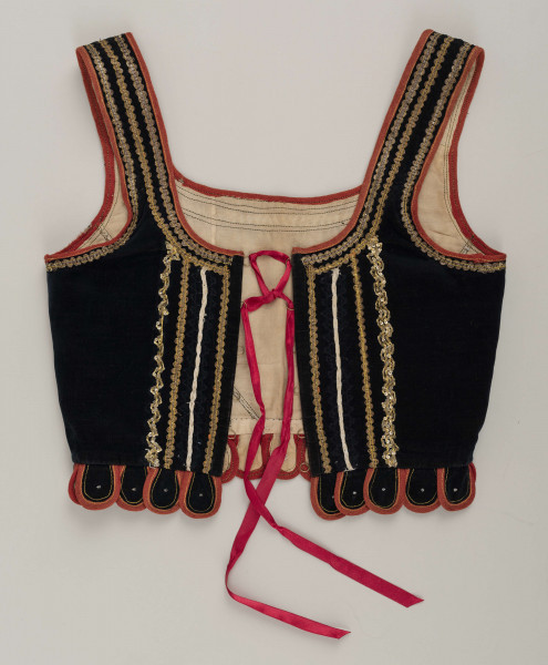 E/9558/ML - Gorset kobiecy z czarnego aksamitu, podszyty białą bawełnianą podszewką. Z przodu rozcięcie usztywnione fiszbinami, zaopatrzone w 10 drucianych haftek do przewlekania wstążeczki. Przód gorsetu ozdobiony złotym szychem, białymi tasiemkami i złotymi drobnymi cekinami naszytymi na nitki w ten sposób, że tworzą ornament w postaci falistej linii. Przód gorsetu zdobiony złotym i srebrnym szychem, klapki brzegi wykroju pachy wykończone czerwoną wełnianą tasiemką. Plecy w miejscach szwów ozdobione żółtą stebnówką i białymi koralikami. Od pasa gorsetu dookoła opadają klapki. Gorset szyty na maszynie.