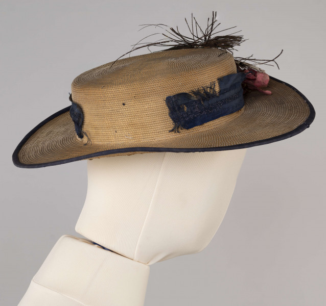 E/560/ML - Kapelusz męski, z cienkiej trzcinki zszywanej nicią. Rondo obszyte niebieską tasiemką, opasane również niebieską (ciemnoniebieską/jasnogranatową) jedwabną wstążką. Kapelusz ozdobiony jest sztucznymi kwiatami białymi i jasnoróżowymi oraz pawimi piórami. Pióra przypiete są metalowo-plastikową broszką.