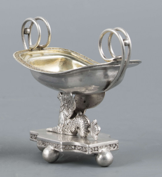 S/Mt/359/ML - Solniczka w kształcie prostokątnej łódki z dwoma wolutowymi uchami z okrągłego drutu, umieszczonej na figurce gryfa, z  podstawą prostokątną o wklęsłych narożach i czterech nóżkach w formie spłaszczonych kulek. Brzeg łódki wywinięty, wnętrze złocone. Brzegi łódki i boki podstawy obiega moletowana taśma z ornamentem roślinnym.