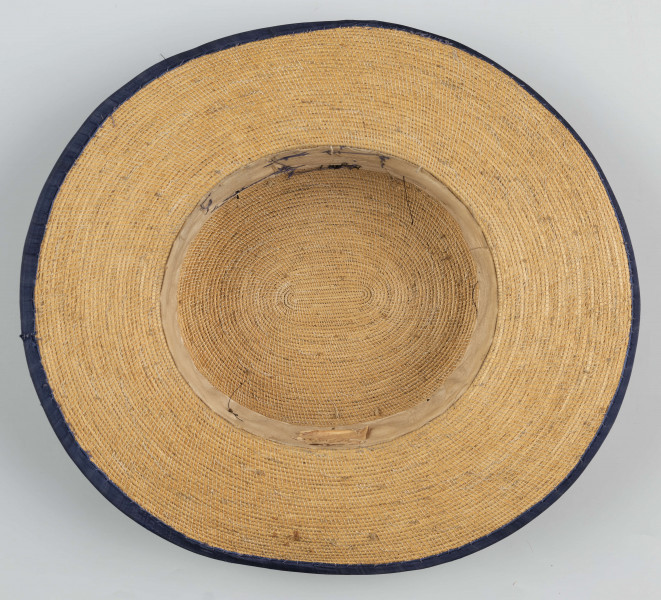 E/560/ML - Kapelusz męski, z cienkiej trzcinki zszywanej nicią. Rondo obszyte niebieską tasiemką, opasane również niebieską (ciemnoniebieską/jasnogranatową) jedwabną wstążką. Kapelusz ozdobiony jest sztucznymi kwiatami białymi i jasnoróżowymi oraz pawimi piórami. Pióra przypiete są metalowo-plastikową broszką.