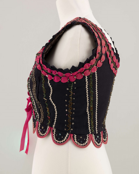 E/7358/ML - Gorset zszyty z 3 kawałków granatowej wełenki, podszyty brązową bawełnianą podszewką. Z przodu rozcięcie usztywnione fiszbinami, zaopatrzone w 10 haftek drucianych do przewlekania wstążeczki. Od pasa gorsetu dookoła opadają klapki. Cały gorset bogato zdobiony amarantowymi tasiemkami, żółtym i srebrnym szychem, białą krepiną, barwnymi koralikami i cekinami. Przód gorsetu, brzegi wykroju i pachy ozdobione dodatkowo granatowymi ząbkami. 