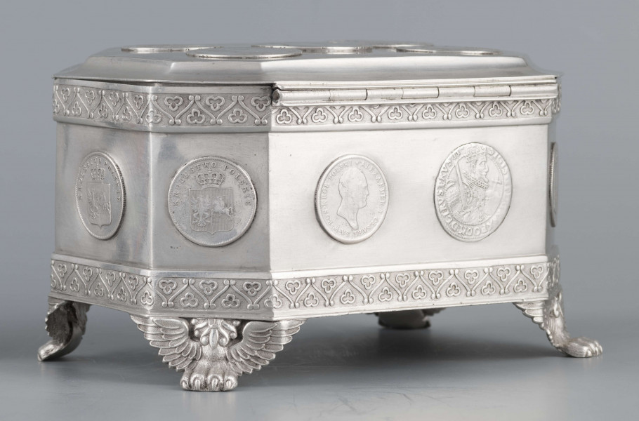 S/Mt/364/ML - Cukiernica skrzynkowa prostokątna ze ściętymi narożami, na czterech nóżkach w formie uskrzydlonych lwich łap, z wieczkiem o wklęsło profilowanym brzegu. Górny i dolny brzeg obiega  moletowana taśma z motywem neogotyckich trójliści, szyld zamka ma formę różanego wieńca pod skrzydatą główką..W ścianki i wieczko wmontowanych jest 15 monet srebrnych:
1 ort Zygmunta III
1 talar pruski, 1818 r.
1 pięciofrankówka, 1811 r.
1 moneta austriacka, 1797 r.
1 moneta saska, 
2 monety polskie St.Augusta Poniatowskiego
1 dwózłotówka Królestwa Polskiego, 1825 r.
1 pięciozłotówka, 1831 r.
6 złotówek, 1831 r.
Na dnie sygnatura wklęsła kursywą:
- K. Rotkiel w Lublinie
i punca z oznaczeniem próby 