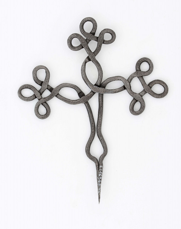 Krzyż żelazny stanowiący zwieńczenie drewnianego krzyża, kowalskiej roboty. Wykonany z pręta o śred. 0,7 cm. Ramiona i wierzchołek krzyża ozdobnie plecione. Szpikulec dł. 6,5 cm.