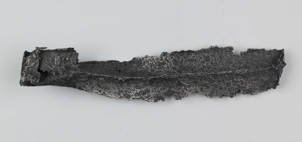 Żelazny grot oszczepu z ostrym żeberkiem środkowym, zgięty rytualnie. Grot stanowi element wyposażenia ciałopalnego pochówku złożonego prawdopodobnie w popielnicy. Odkrycia dokonano w 1959 roku przypadkowo w czasie prac rolnych. Oprócz trzech grotów, pochówkowi wojownika towarzyszyły: żelazne umbo, dwie ostrogi, tok brązowy (prawdopodobnie okucie drzewca oszczepu) a także żelazny miecz z miedziana inkrustacją wyobrażającą rzymskich bogów. Pochowany na terenie obecnej wsi Podlodów wojownik należał do elity wojskowej przedstawicieli kultury przeworskiej.