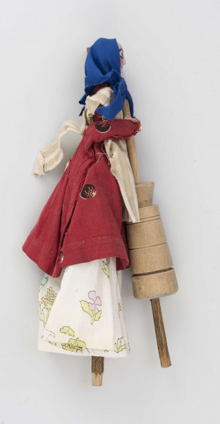 Kukiełka z szopki przedstawiająca czarownicę robiącą masło w maselnicy. Postać ubrana w jasną długą spódnicę, czerwony w koła kaftan i fartuch w szare pionowe pasy na jasnym tle. Na głowie ma granatową jedwabną chustkę wiązaną pod brodą. Twarz słabo modelowana, nos wystający, oczy namalowane farbą. Maselnica toczona, przyczepiona drutem do kukiełki.