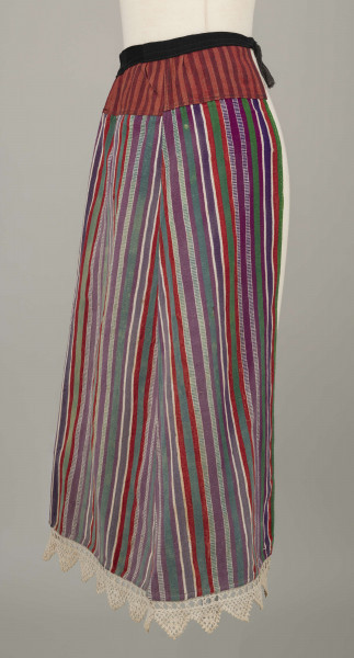 E/8132/ML - Fartuch wełniany szyty maszynowo. Wykonany z materiału samodziałowego tkanego w pionowe paski, w kolorach: czerwonym, białym, szafirowym, bordowym, zielonym. Uszyta z prostokątnego kawałka materiału, do którego doszyto z obu stron kliny. U góry fartuszek przedłużony przez wszycie kawałka materiału szer. 7 cm, w pasy czerwono-granatowe. W talii wszyty w pasek z czarnego materiału, szer. 2 cm, zakończony troczkami. Dół fartuszka zdobi biała koronka szer. 5 cm.