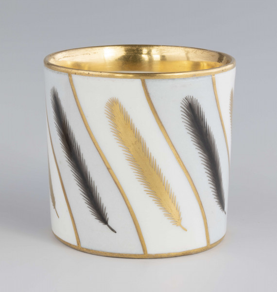 Filiżanka i spodek malowane w piórka. Filiżanka cylindryczna, ucho łamane z listkami w osadzeniach. Brzeg złocony obustronnie, złotem  konturowane ucho, złote kontury dzielą powierzchnię na ukośne pasy na przemian białe i jasnoszare. W nich malowane odpowiednio złote i czarne pióra.