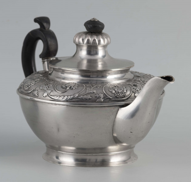 S/Mt/542/ML - Korpus w kształcie spłaszczonej kuli, na niskiej stopie, wylew w formie dzióbka, ucho drewniane malowane na czarno, osadzone w dwóch tulejkach, pokrywka na zawiasie, płaska, w części środkowej wypiętrzona i ozdobiona puklowaną rozetą.Górna część brzuśca ozdobiona sztancowaną taśmą z wzorem spiralnie zwinietej wici roślinnej z kwiatami, uchwyt przykrywki zakończony drewnianą rozetką.  
