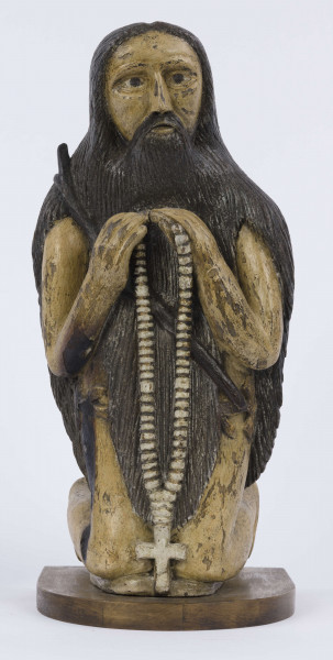 E/6353/ML - Figura drewniana, zakomponowana na postumencie, polichromowana. Postać klęcząca przedstawia św. Onufrego. Twarz płaska, trójkątna, głęboko osadzone oczy, czarne źrenice. Szerokie łuki brwiowe łączą się z wydatnym prostym nosem. Wystające kości jarzmowe. Małe usta otacza bujny zarost: broda i wąsy łączące się z bujnymi włosami, otaczającymi całą postać – na podobieństwo płaszcza. Ręce zgięte w łokciach, uniesione ku górze, łączą się końcami palców w geście modlitwy. W nieproporcjonalnie dużych dłoniach biały różaniec zakończony dużym krzyżem. Nogi masywne z dużymi płaskimi stopami. Prawą dłonią podtrzymuje sękaty kostur. W partii pasa przepasany powrozem z węzłami. 