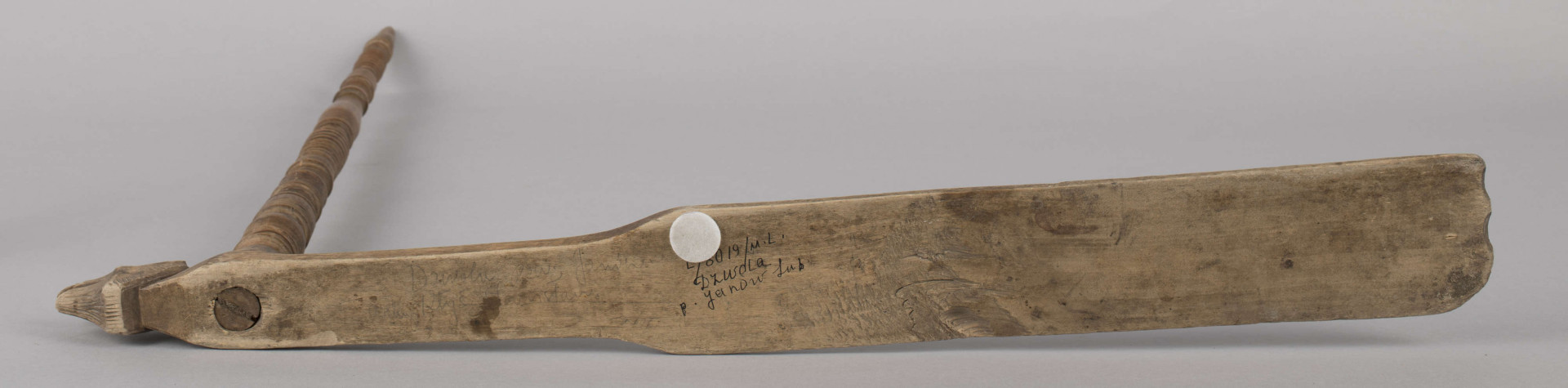 E/8019/ML - Przęślica krężołkowa z drewna brzozowego składa się z cienkiej deski – przysiadki, w której przy jednym końcu osadzona jest pionowa beleczka zw. piórem, z nasadzonym od góry ruchomym krężlem w kształcie wydłużonego stożka. Deseczka przysiadki o kształcie prostokątnym lekko zwężającym, od ok. połowy swej długości w kierunku pióra, i zakończonym głową zwierzęcą – psa. Pióro i krężel ozdobnie toczone. Krężel ma kształt stożka z rozszerzoną podstawą, w której znajduje się stożkowaty otwór, do osadzenia krężla na piórze. Dekoracja snycerska ryta znajduje się na zwężającej części przysiadki. Część zdobiona oddzielona małym stopniem. Grubość przysiadki w części zdobionej 2x większa od pozostałej części. Dekoracja snycerska tworzy ornament roślinny w postaci trzech ulistnionych drobno gałązek. Na zakończeniu ornamentu znajdują się ryte liście oraz inicjały „K.F.”. Głowa psa zdobiona drobnymi nacięcia. Zakończenie przysiadki wycięte faliście.