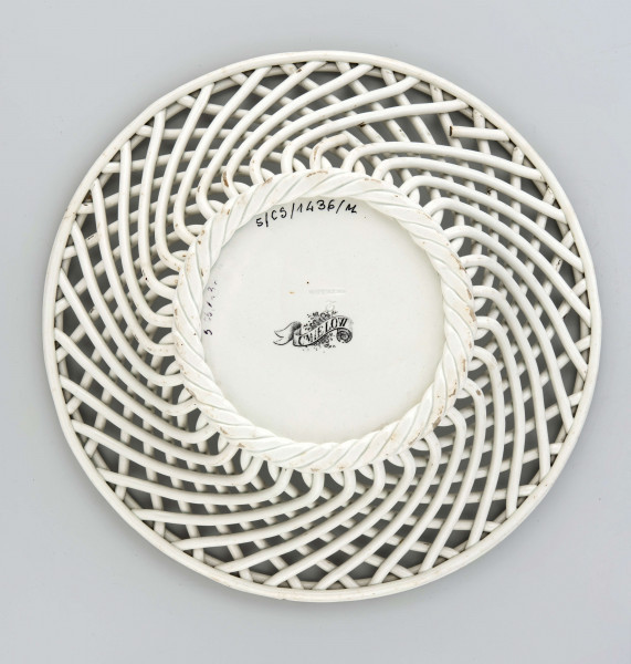 Koszyczek okrągły, szkliwiony, biały, dno gładkie, kołnierz ażurowy z okrągłych ukośnie wygiętych wałków ułożonych  w dwóch warstwach przciwlegle, zebranych potrójnym wałkiem tworzącym krawędź .W lustrze dekoracja w technice stalorytu w  kolorze czarnym: scenka rodzajowa w pejzażu o cechach południowych (palmy) z architekturą, w centrum dwukołowy wóz ciągnięty przez woły i postacie ludzi.
 
