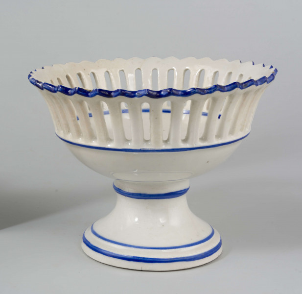 Patera w formie kielicha na okrągłej nóżce, czara w górnej części ażurowa z pionowych pasków zamkniętych ząbkowanym brzegiem. Pokryta białym szkliwem, profile i krawędzie podkreślone paskami w kolorze błękitu kobaltowego.
 