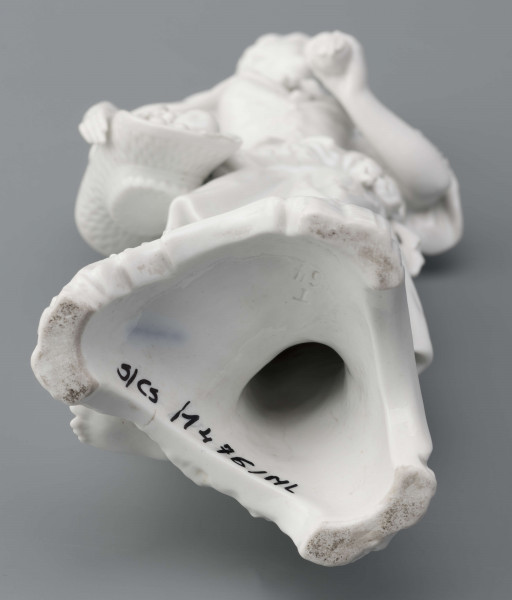 S/CS/1476/ML - Figurka z białej szkliwionej porcelany przedstawiajaca  kobietę stojacąna niskim trójdzielnym cokole. Kobieta ma długie włosy upięte w koczek, ubrana jest w fałdzistą spódnicę przewiązaną poniżej bioder, gorset z baskinką i głębokim dekoltem, bluzkę z bufiastmi rękawami do łokci, na ramionach zawiazaną chustkę, ma bose stopy. Prawą ręką trzyma płaski kosz z owocami oparty na biodrze, w lewej ugiętej ma jabłko. 
  