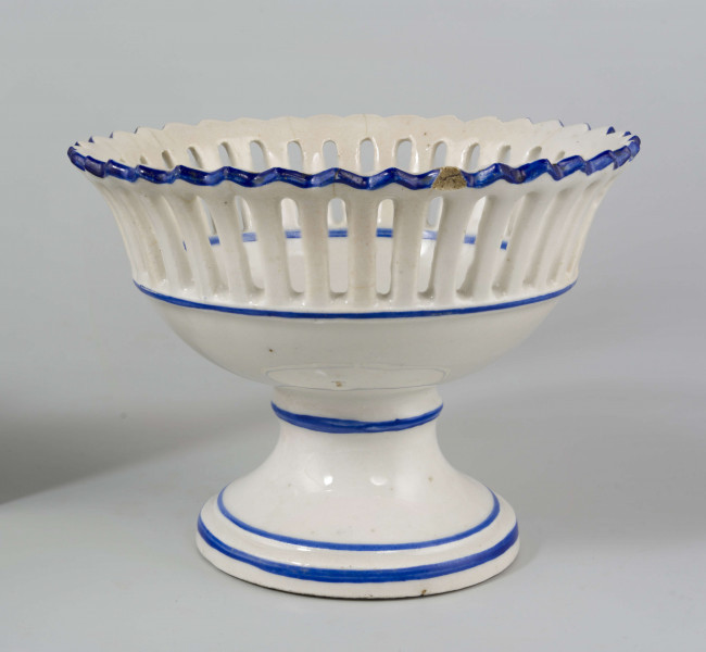 Patera w formie kielicha na okrągłej nóżce, czara w górnej części ażurowa z pionowych pasków zamkniętych ząbkowanym brzegiem. Pokryta białym szkliwem, profile i krawędzie podkreślone paskami w kolorze błękitu kobaltowego.
 