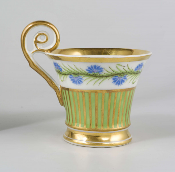 Filiżanka koniczna na pierścieniowatej stopie, ucho w formie pastorału z wolutą wyciągniętą ponad krawędź wylewu. Dekoracja malowana i złocona pokrywa całą powierzchnię: wokół wylewu fryz w postaci girlandy błękitnych chabrów z zielonymi listkami na białym tle, poniżej wzór pionowych pasków naprzemian złotych i zielonych; stopa, wylew (obustronnie) i ucho podkreślone złotymi paskami. 