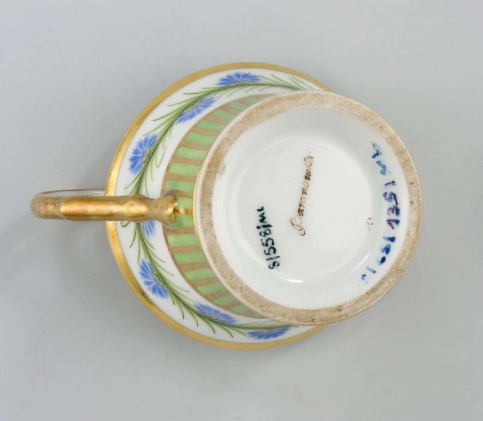 Filiżanka koniczna na pierścieniowatej stopie, ucho w formie pastorału z wolutą wyciągniętą ponad krawędź wylewu. Dekoracja malowana i złocona pokrywa całą powierzchnię: wokół wylewu fryz w postaci girlandy błękitnych chabrów z zielonymi listkami na białym tle, poniżej wzór pionowych pasków naprzemian złotych i zielonych; stopa, wylew (obustronnie) i ucho podkreślone złotymi paskami. 