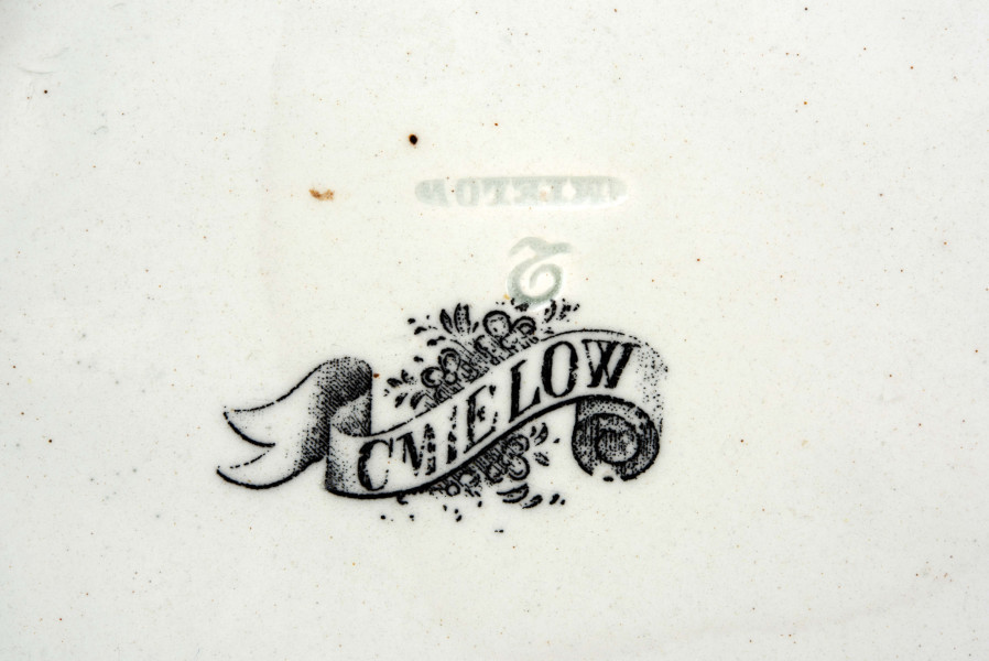 Koszyczek okrągły, szkliwiony, biały, dno gładkie, kołnierz ażurowy z okrągłych ukośnie wygiętych wałków ułożonych  w dwóch warstwach przciwlegle, zebranych potrójnym wałkiem tworzącym krawędź .W lustrze dekoracja w technice stalorytu w  kolorze czarnym: scenka rodzajowa w pejzażu o cechach południowych (palmy) z architekturą, w centrum dwukołowy wóz ciągnięty przez woły i postacie ludzi.
 
