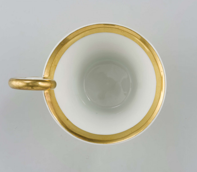 Filiżanka koniczna na pierścieniowatej stopie, ucho w formie pastorału z wolutą wyciągniętą ponad krawędź wylewu. Dekoracja malowana i złocona pokrywa całą powierzchnię: wokół wylewu fryz w postaci girlandy błękitnych chabrów z zielonymi listkami na białym tle, poniżej wzór pionowych pasków naprzemian złotych i zielonych; stopa, wylew (obustronnie) i ucho podkreślone złotymi paskami. 