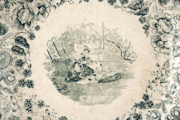 Talerz deserowy, okrągły, gładki, pokryty szkliwem białym, na nim dekoracja  drukowana w kolorze zieleni chromowej: w lustrze scena rodzajowa - kobieta z bawiącym sie dzieckiem; na kołnierzu szeroka bordiura z motywem ząbkowanej koronki, ornamentów wolutowych i kwiatów. 
 