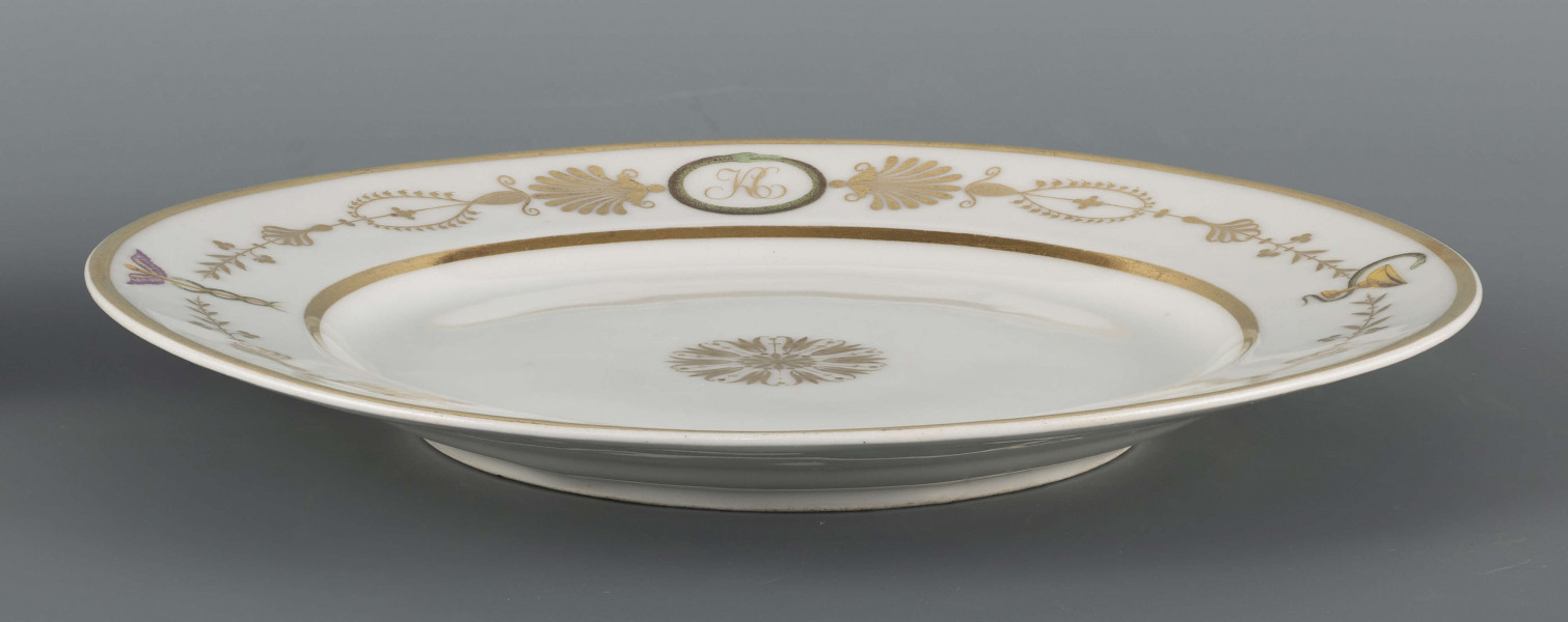 S/CS/1594/ML - Talerz płytki, okrągły, gładki. Na kołnierzu fryz z ornamentu palmetowego przeplecionego barwnymi elementami: kaduceuszem, wężem Eskulapa, monogramem 