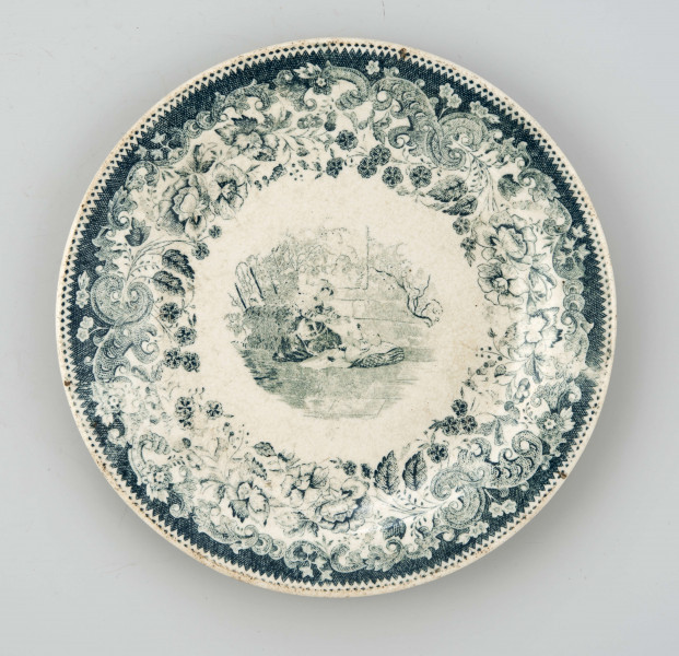 Talerz deserowy, okrągły, gładki, pokryty szkliwem białym, na nim dekoracja  drukowana w kolorze zieleni chromowej: w lustrze scena rodzajowa - kobieta z bawiącym sie dzieckiem; na kołnierzu szeroka bordiura z motywem ząbkowanej koronki, ornamentów wolutowych i kwiatów. 
 