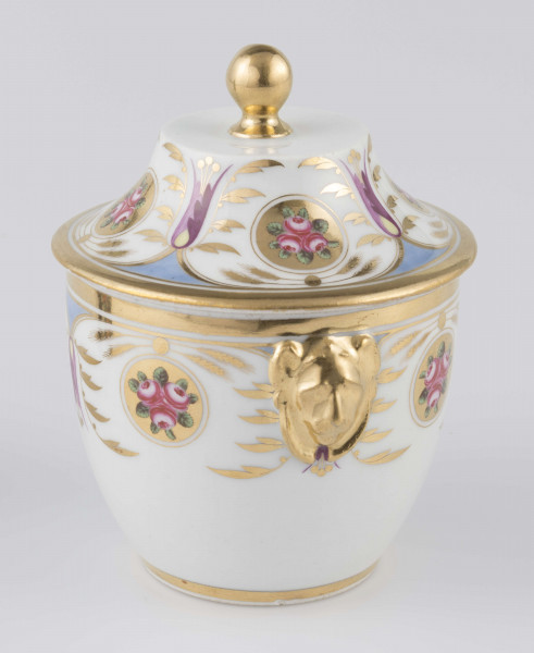 S/CS/1388/2/ML - Cukiernica półowoidalna z uchwytami w formie półplastycznych lwich głów i pokrywką w kształcie ściętego stożka o wklęsłych bokach z kulistym uchwytem. Dekoracja malowana wielobarwnie i złocona: brzusiec i boki pokrywki obiega szeroki fryz złożony ze stylizowanych ornamentów roślinnych (gałązek, kłosów zbóż i liści akantu)  połączonych z owalnymi medalionami, w których są trzy różowe różyczki z zielonymi listkami na złotym tle.Uchwyty malowane złotem,  złotym konturem obwiedzione wszystkie krawędzie.
 