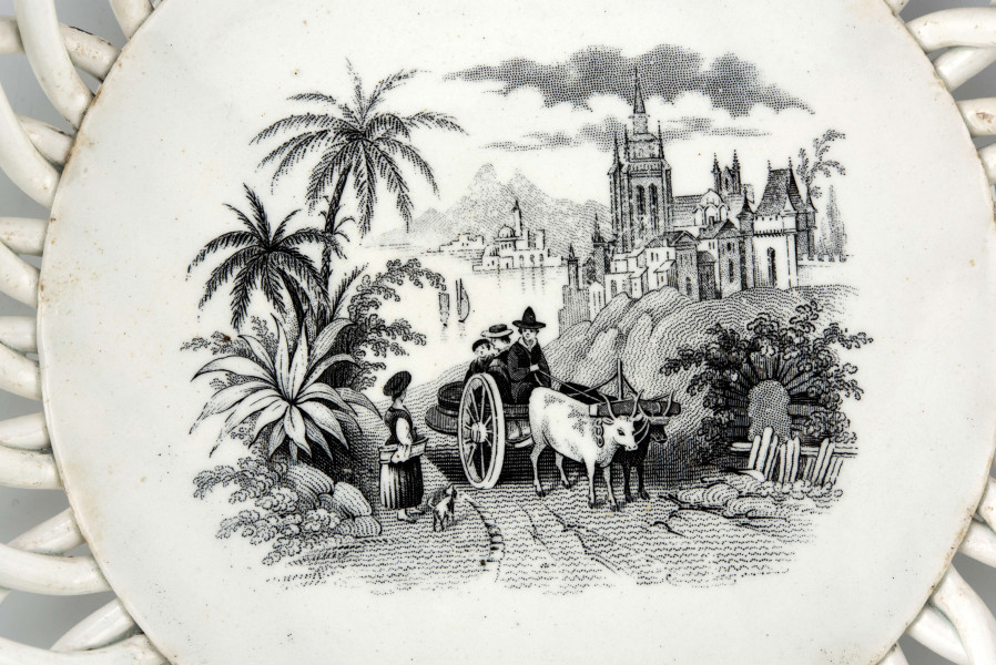 Koszyczek okrągły, szkliwiony, biały, dno gładkie, kołnierz ażurowy z okrągłych ukośnie wygiętych wałków ułożonych  w dwóch warstwach przciwlegle, zebranych potrójnym wałkiem tworzącym krawędź .W lustrze dekoracja w technice stalorytu w  kolorze czarnym: scenka rodzajowa w pejzażu o cechach południowych (palmy) z architekturą, w centrum dwukołowy wóz ciągnięty przez woły i postacie ludzi.
 
