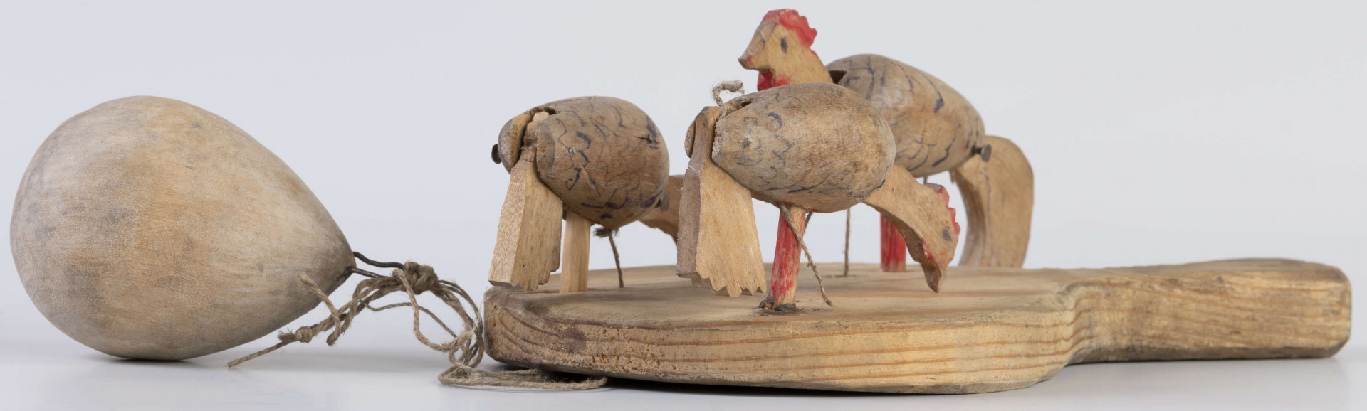 E/17030/ML - Zabawka drewniana przedstawiająca trzy kurki przymocowane do owalnej deseczki z uchwytem. Kurki mają ruchome głowy i ogony, które poruszają się za pomocą sznurków przeciągniętych przez deseczkę i przywiązanych do drewnianej kuli. Deseczka wykonana z drewna sosnowego, kurki z drewna lipowego. Głowy i nogi kurek pomalowane są czerwoną farbą, tułowia zdobi ornament geometryczny w postaci zygzakowatych linii wykonanych brązową farbą.