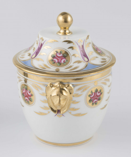 S/CS/1388/2/ML - Cukiernica półowoidalna z uchwytami w formie półplastycznych lwich głów i pokrywką w kształcie ściętego stożka o wklęsłych bokach z kulistym uchwytem. Dekoracja malowana wielobarwnie i złocona: brzusiec i boki pokrywki obiega szeroki fryz złożony ze stylizowanych ornamentów roślinnych (gałązek, kłosów zbóż i liści akantu)  połączonych z owalnymi medalionami, w których są trzy różowe różyczki z zielonymi listkami na złotym tle.Uchwyty malowane złotem,  złotym konturem obwiedzione wszystkie krawędzie.
 