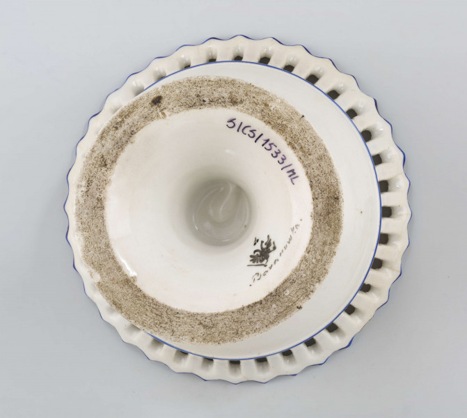 Patera w formie kielicha na okrągłej nóżce, czara w górnej części ażurowa z pionowych pasków zamkniętych ząbkowanym brzegiem. Pokryta białym szkliwem, profile i krawędzie podkreślone paskami w kolorze błękitu kobaltowego.
 