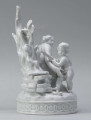 S/CS/1456/ML - Grupa figuralna z białej szkliwionej porcelany. Cokół owalny, wysklepiony z ornamentem opaskowym złożonym ze splecionych owali z rozetkami. Na nim siedzi naga kobieta (Venus) przed płonącym stosem o wysokich płomieniach i wyciągniętą w bok prawą ręką powstrzymuje nagie dziecko (Amorek) zapalające od ognia pochodnię, lewą ręką siega do ażurowego naczynia z ogniem. Za nią na powierzchni cokołu drobne roślinki.

