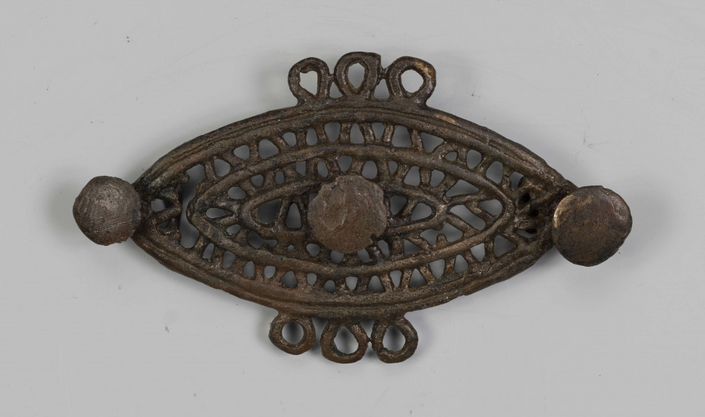 1396/A/ML/2 - Ażurowa fibula z brązu będąca elementem dwuczęściowej ozdoby. Na ornament składają się trzy koncentryczne listwy soczewkowate połączone ze sobą naprzemianlegle skośnymi, krótkimi poprzeczkami, tworzącymi trójkąty. Na dłuższej osi tarczki umieszczone są trzy symetrycznie guzki. Po środku dłuższych boków tarczki, naprzeciw siebie znajdują się potrójne uszka. 
