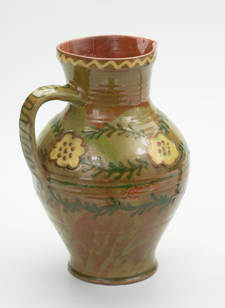 Gliniany dzban zdobiony motywem kwiatowym - Ujęcie skosem w lewą stronę. Dzban gliniany. Brzusiec mocno uwypuklony przechodzi w szeroką nieco rozchyloną szyjkę z małym dziobkiem. Ucho taśmowe wzdłużne. Wewnątrz glazura brązowa, na zewnątrz zielona. Dookolny ornament jodełkowy na brzuścu. Krawędź szyjki ozdobiona linią falistą. Ornament wykonany emalią zieloną i żółtą.