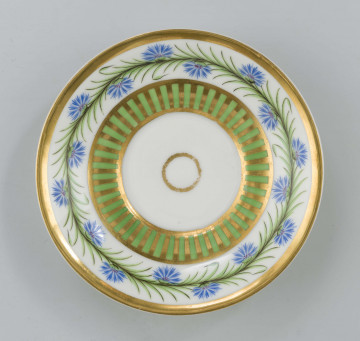Spodek miseczkowaty na pierścieniowej podstawie, wgłębiony, dekoracja malowana wielobarwnie i złocona: pośrodku dna złote kółko,  wokół dna wzór koncentrycznych pasków na przemian złotych i zielonych, na kołnierzu  fryz w postaci girlandy błękitnych chabrów z zielonymi listkami na białym tle, krawędź obwiedziona złotym paskiem.     