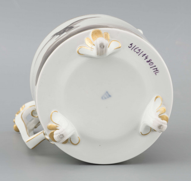 S/CS/1480/ML - Dzbanek cylindryczny, na trzech nóżkach w kształcie lwich łap, szyja w formie zwężonego pierścienia, wylew z dziobkiem, ucho dołem prostokątnie załamane, górą wygięte, rozdwojone, z reliefowymi liśćmi w osadzeniach. Przykrywa w formie wąskiego, konicznego pierścienia, z uchwytem w kształcie szyszki. Dekoracja drukowana en grisaille w kolorze czarnym. Po obu stronach brzuśca sceny alegoryczne: lew prowadzony na pasku przez amorka, powyżej napis 