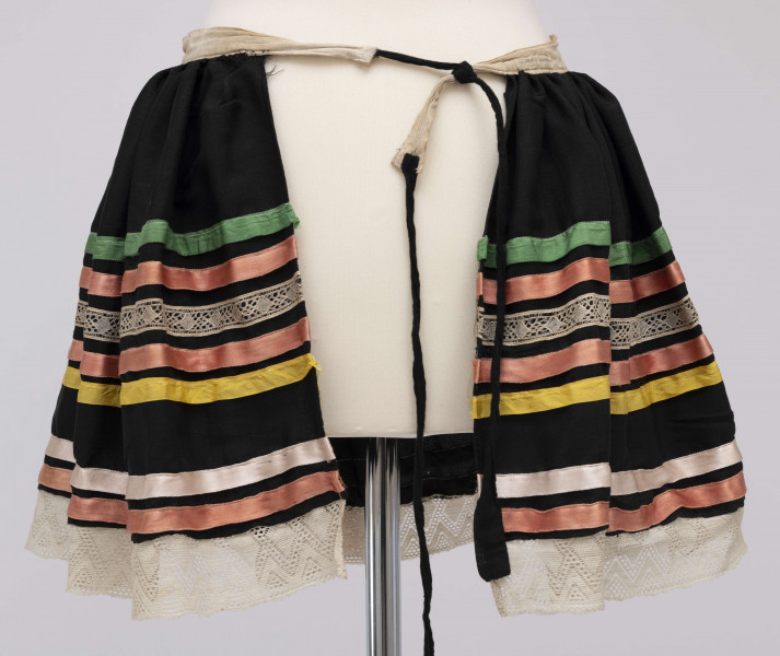 E/10495/ML - Fartuszek z cienkiej wełenki w kolorze czarnym, wszyty w płócienną oszewkę i wiązany na troczki. Suto marszczony. W odległości 13 cm od pasa naszyte wstążki (szer. 1,5 cm) i koronki (szer. 2 cm) w kolorach: zielonym, czerwonym, koronka biała, czerwonym i żółtym. Dół wykończony białą koronką (szer. 4,5 cm). Powyżej naszyte 2 wstążki: czerwona i różowa.