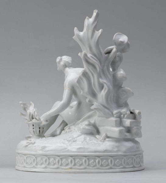 S/CS/1456/ML - Grupa figuralna z białej szkliwionej porcelany. Cokół owalny, wysklepiony z ornamentem opaskowym złożonym ze splecionych owali z rozetkami. Na nim siedzi naga kobieta (Venus) przed płonącym stosem o wysokich płomieniach i wyciągniętą w bok prawą ręką powstrzymuje nagie dziecko (Amorek) zapalające od ognia pochodnię, lewą ręką siega do ażurowego naczynia z ogniem. Za nią na powierzchni cokołu drobne roślinki.
