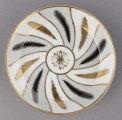 S/CS/1678/2/ML - Spodek okrągły, konicznie wgłębiony, brzeg złocony, złote kontury dzielą powierzchnię na ukośne pasy na przemian białe i jasnoszare. W nich malowane odpowiednio złote i czarne pióra w lustrze pośrodku złota rozeta.
 