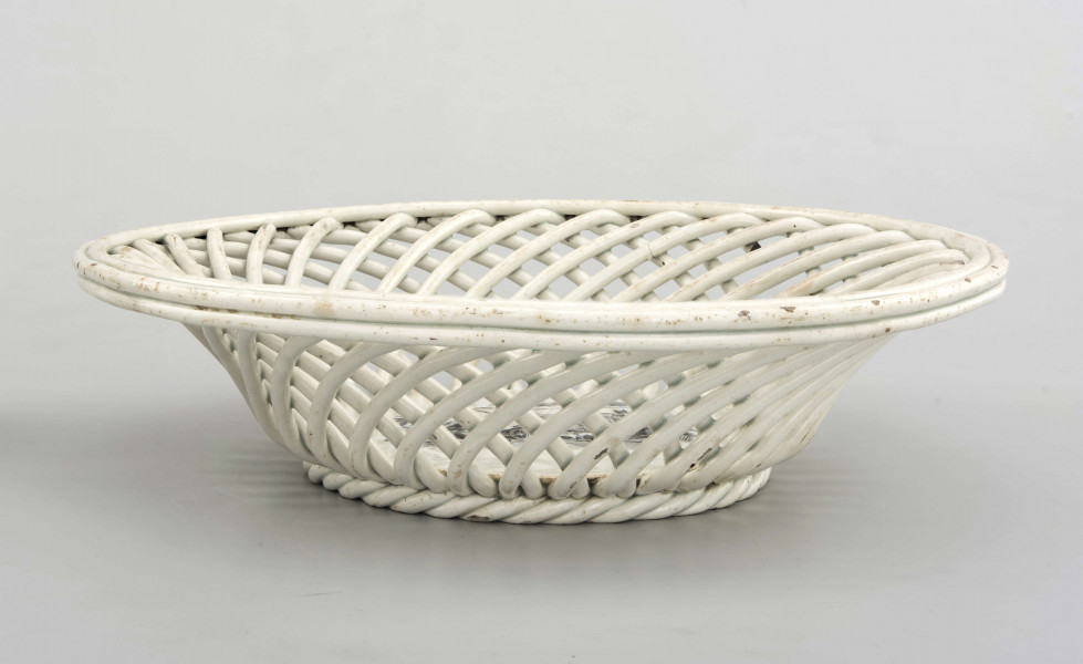 Koszyczek okrągły, szkliwiony, biały, dno gładkie, kołnierz ażurowy z okrągłych ukośnie wygiętych wałków ułożonych  w dwóch warstwach przciwlegle, zebranych potrójnym wałkiem tworzącym krawędź .W lustrze dekoracja w technice stalorytu w  kolorze czarnym: scenka rodzajowa w pejzażu o cechach południowych (palmy) z architekturą, w centrum dwukołowy wóz ciągnięty przez woły i postacie ludzi.
 
