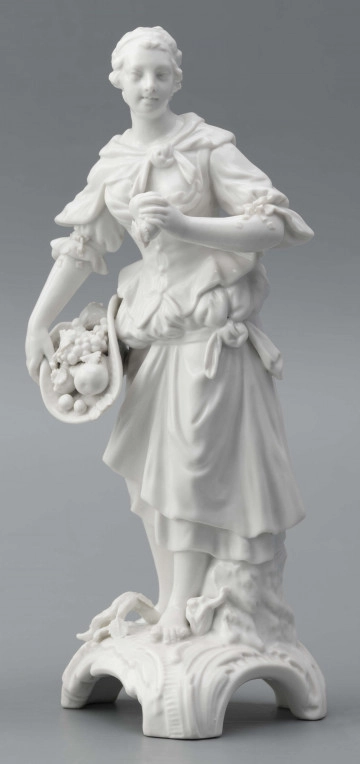 S/CS/1476/ML - Figurka z białej szkliwionej porcelany przedstawiajaca  kobietę stojacąna niskim trójdzielnym cokole. Kobieta ma długie włosy upięte w koczek, ubrana jest w fałdzistą spódnicę przewiązaną poniżej bioder, gorset z baskinką i głębokim dekoltem, bluzkę z bufiastmi rękawami do łokci, na ramionach zawiazaną chustkę, ma bose stopy. Prawą ręką trzyma płaski kosz z owocami oparty na biodrze, w lewej ugiętej ma jabłko. 
  