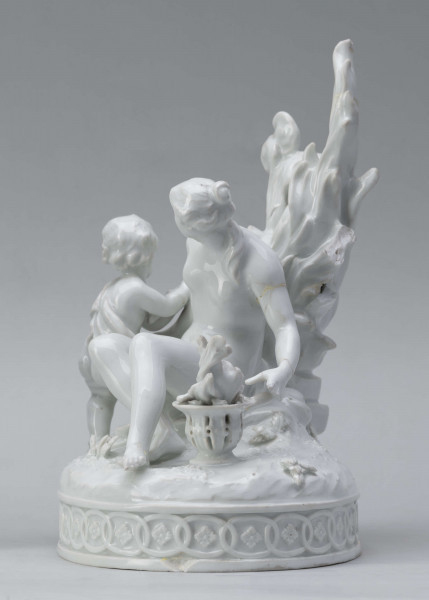 S/CS/1456/ML - Grupa figuralna z białej szkliwionej porcelany. Cokół owalny, wysklepiony z ornamentem opaskowym złożonym ze splecionych owali z rozetkami. Na nim siedzi naga kobieta (Venus) przed płonącym stosem o wysokich płomieniach i wyciągniętą w bok prawą ręką powstrzymuje nagie dziecko (Amorek) zapalające od ognia pochodnię, lewą ręką siega do ażurowego naczynia z ogniem. Za nią na powierzchni cokołu drobne roślinki.
