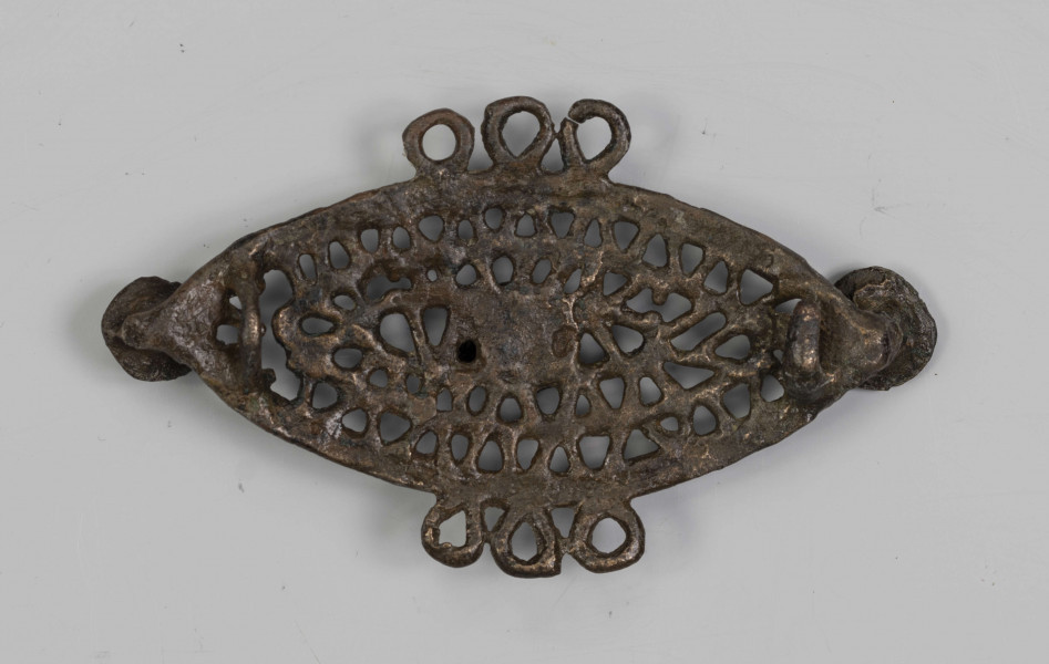 1396/A/ML/2 - Ażurowa fibula z brązu będąca elementem dwuczęściowej ozdoby. Na ornament składają się trzy koncentryczne listwy soczewkowate połączone ze sobą naprzemianlegle skośnymi, krótkimi poprzeczkami, tworzącymi trójkąty. Na dłuższej osi tarczki umieszczone są trzy symetrycznie guzki. Po środku dłuższych boków tarczki, naprzeciw siebie znajdują się potrójne uszka. 