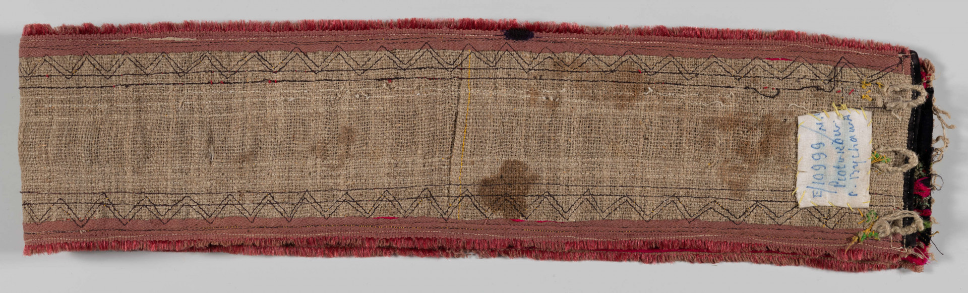 E/10999/ML - Pas męski szyty maszynowo z grubego lnianego płótna. Obszyty szczoteczką w kolorze czerwonym oraz cienką wełną w kolorze amarantowym i taśmą srebrzystą. Na środek naszyty jest czarny aksamit wycięty „w okienka” – ażurowy szlak z rombów. Pod okienka podłożone taśmy biało-czerwono-zielone ze srebrna nitką oraz amarantowe i zielone. Zapinany na 3 ciemne guziki i pętelki ręcznie wykonane z nici. 